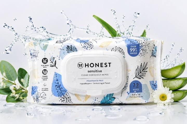 A package of The Honest Company Sensitive Clean Conscious Wipes with a white plastic flip-top lid, featured against a splash of water, green leaves, and sliced aloe vera.